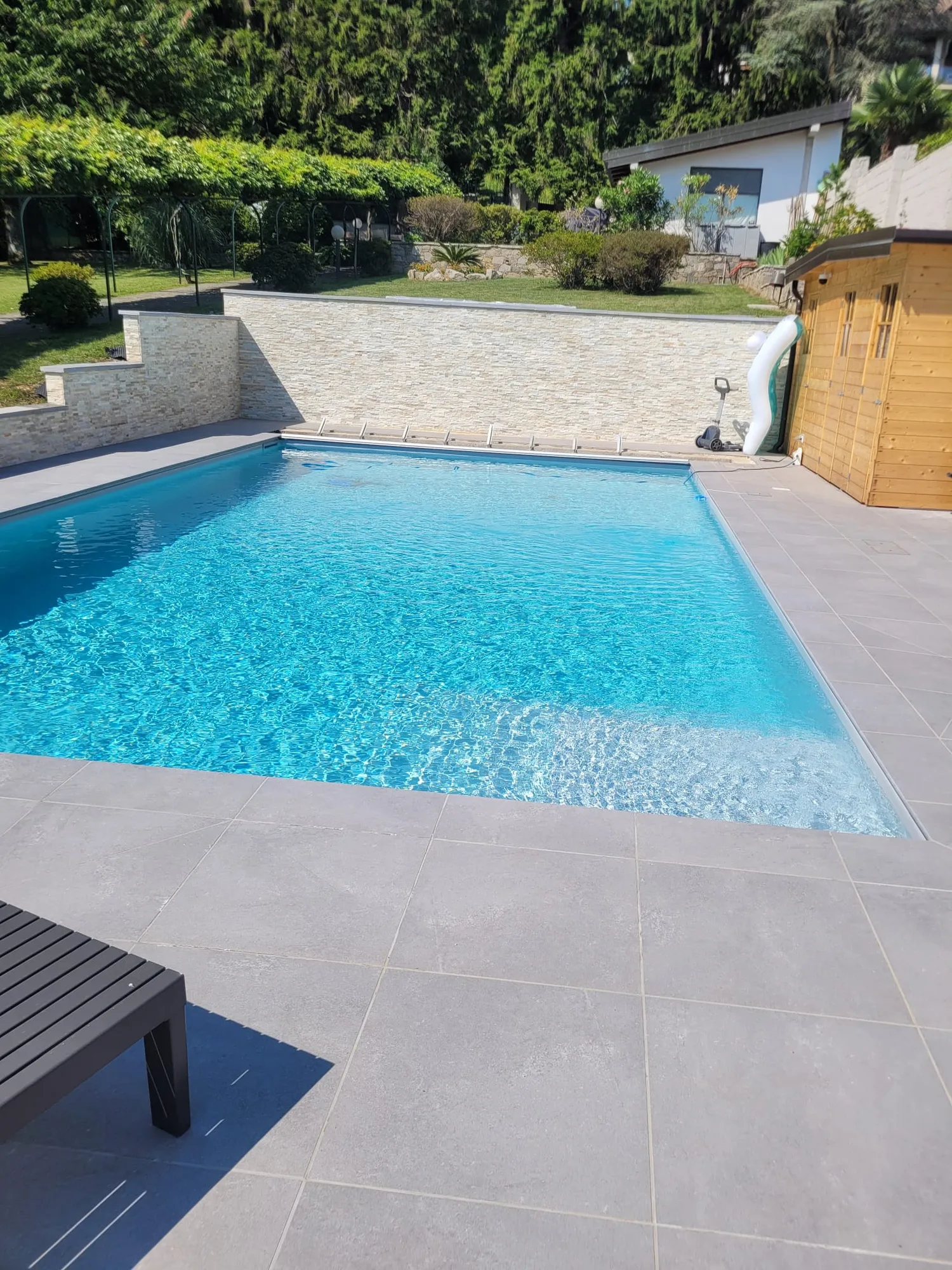 Piscina piena d'acqua con il bordo in gres grigio finito e il giardino intorno